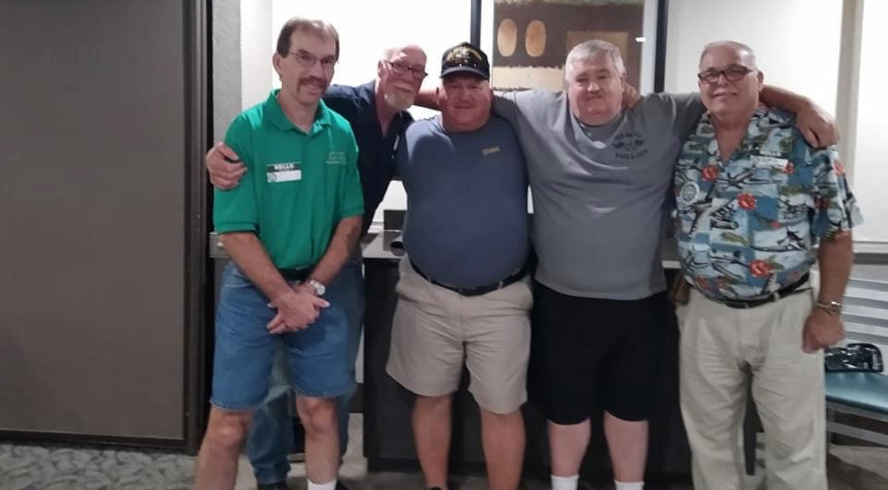 Four men standing in a kitchen with one man holding his arm up.