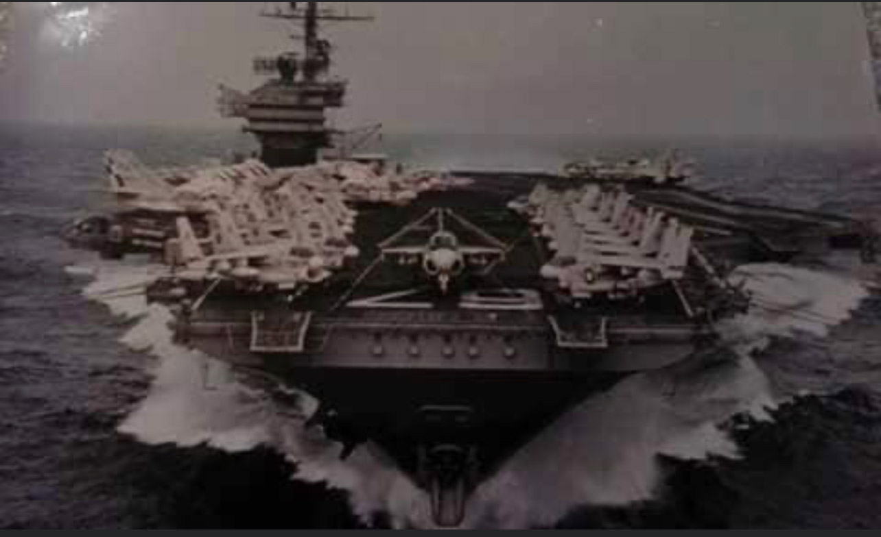 A large aircraft carrier in the ocean with waves crashing behind it.